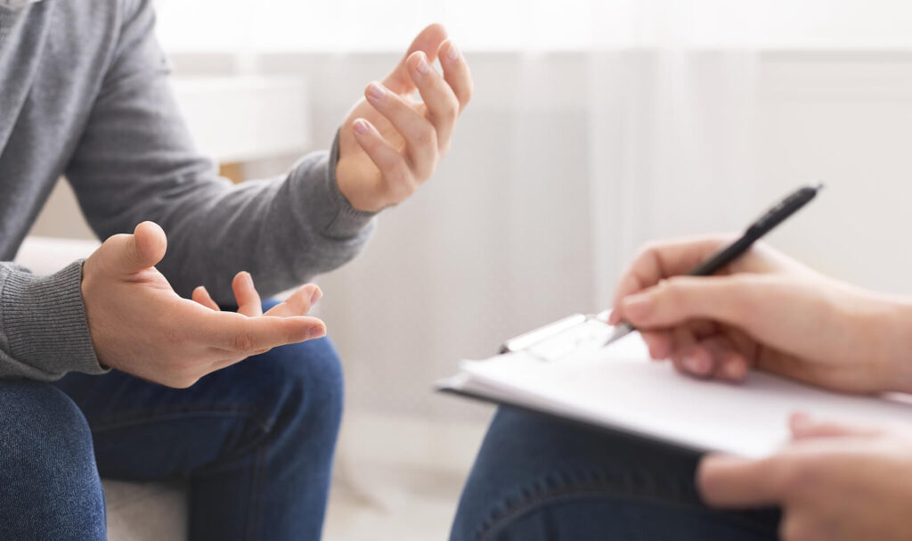 A close up of a person gesturing with their hands while a person with a clipboard takes notes. This could represent the support anxiety treatment in American Fork, UT can offer in addressing post pandemic anxiety. Search for teenage counseling utah or teenage anxiety treatment in American Fork, UT today. 
