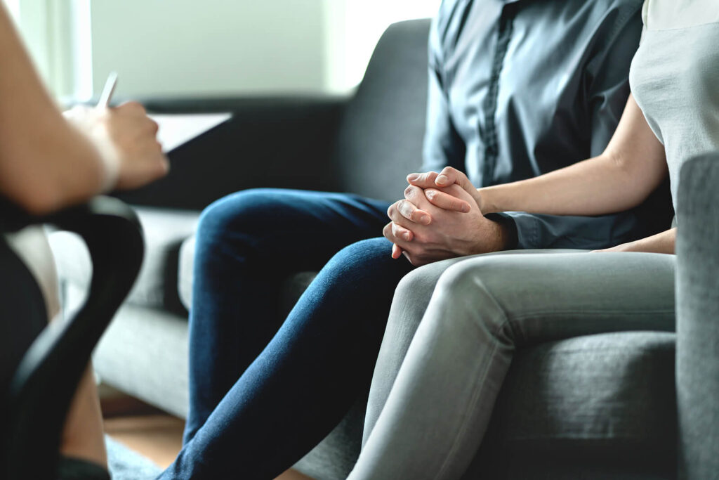 A couple holds hands while sitting on a couch across from a person with a clipboard taking notes. This could represent the support a couples therapist in American Fork, UT can offer. Learn more about couples counseling in American Fork, UT by searching for couples communication therapy American Fork, UT today.
