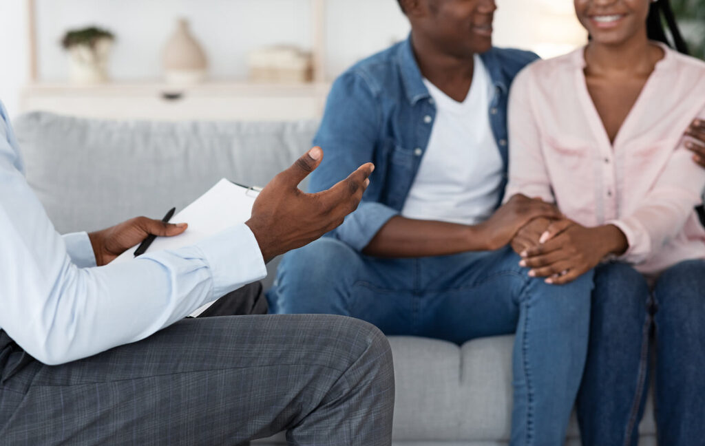 A close up of a couple holding hands while sitting across from a woman writing notes on a notepad. This could symbolize the support couples counseling in American Fork, UT can offer. Search for couples therapy in American Fork, UT to learn more about couples communication therapy and other services. 
