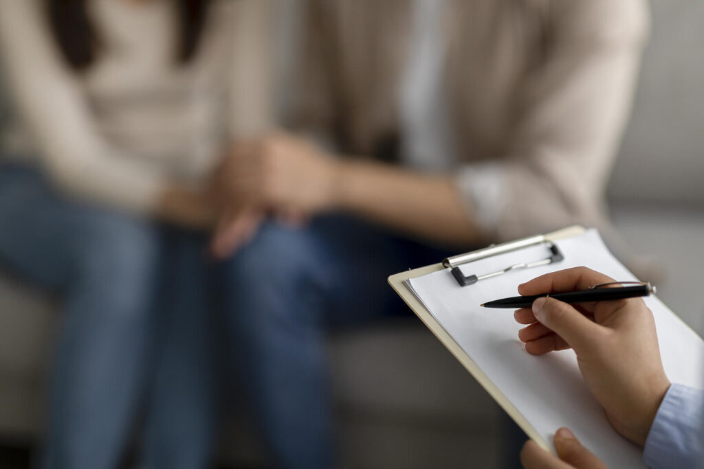 A close up of a person taking notes on a clipboard while sitting across from a couple holding hands. Learn more about how a therapist for couples can provide support with betrayal trauma therapy in American Fork, UT and other services. Search for couples therapy in American Fork, UT for more info.