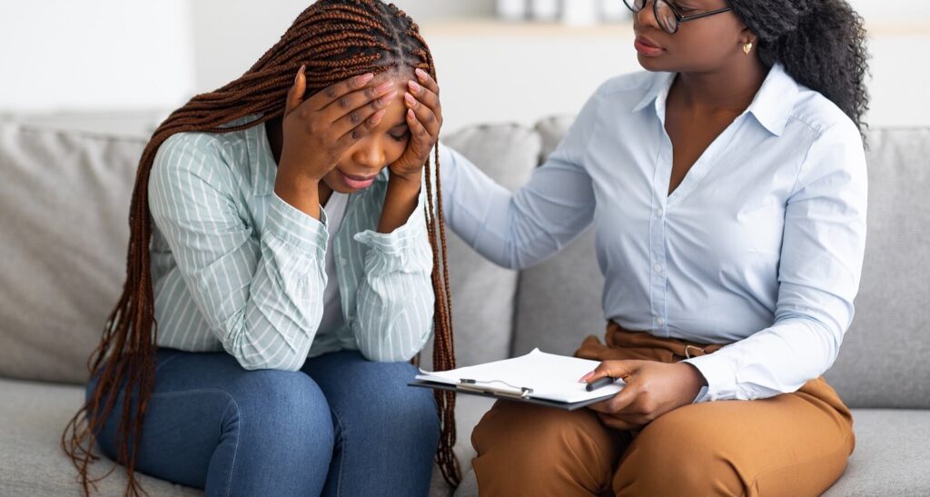 A woman holding both sides of her head receives a supportive hand from a woman with a clipboard. This could represent the support depression therapists in American Fork, UT can provide through therapy for depression in American Fork, UT. Search for teenage depression treatment near me and other services today. 
