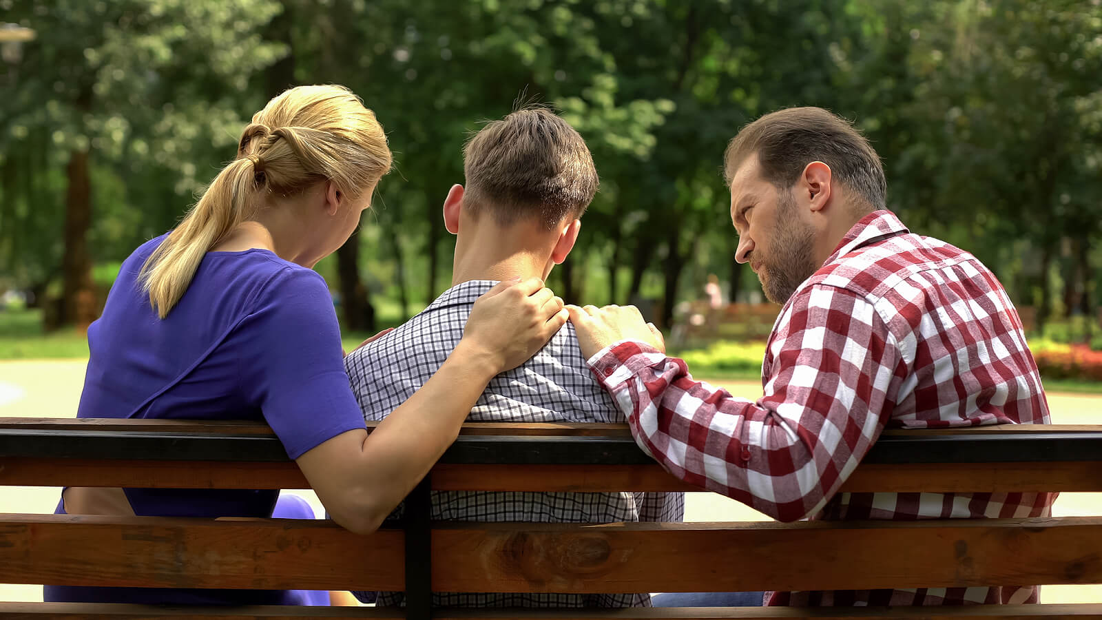 A young man sits on a park bench with a woman and a man on either side, both placing comforting hands on his shoulders. This could represent the support offered for teens from parents and therapy for teens in American Fork, UT. Search for a family therapist American Fork, UT and how therapists for teens American Fork, UT can help.