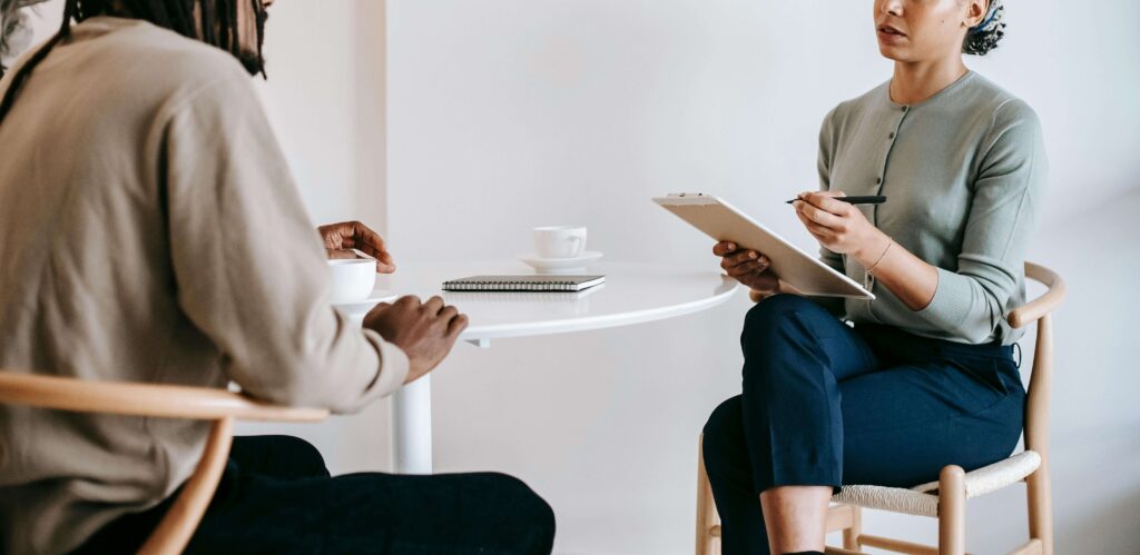 A specialist, holding a clipboard, attentively listens to a client seated across from her at a small white table. This could represent the support a trauma therapist in American Fork, UT can offer for clients. Search for betrayal trauma therapy in American Fork, UT to learn more. 
